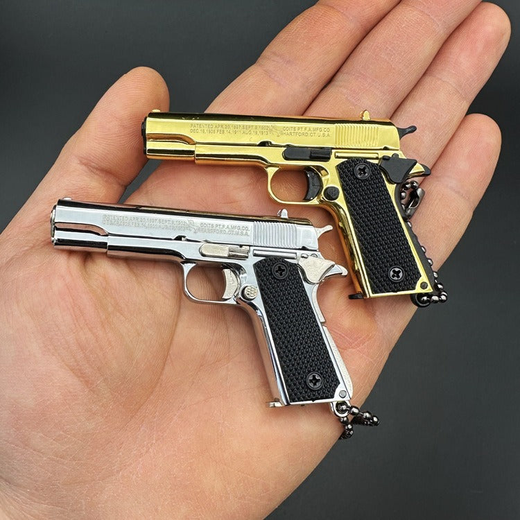 Two miniature gun keychains, one gold and one silver, held in a hand against a dark background.