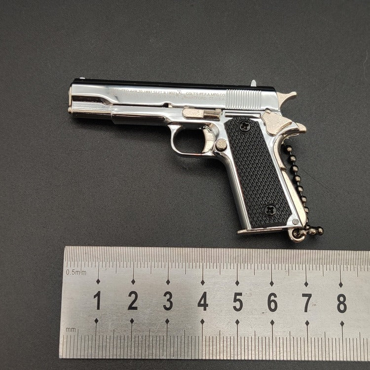 Metallic gun-shaped keychain with black grip on a gray background, accompanied by a ruler for scale.